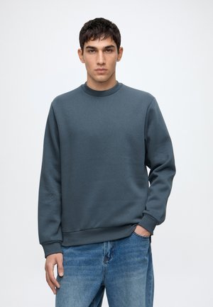 Junger Mann mit kurzen dunklen Haaren, der einen schlichten dunkelgrauen Sweatshirt und blaue Jeans trägt, steht mit einer Hand in der Tasche.