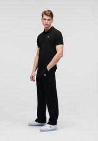 Polo nero a maniche corte con un piccolo logo, abbinato a pantaloni della tuta neri con un logo coordinato. Il modello indossa scarpe da ginnastica bianche.