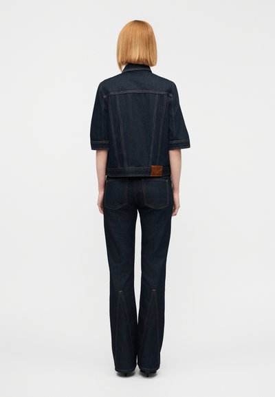 Μαύρο τζιν μπουφάν με κοντό σχέδιο και κοντά μανίκια, συνδυασμένο με φαρδιά μαύρα τζιν παντελόνια. Διαθέτει καφέ δερμάτινο μπάλωμα στο πίσω μέρος.