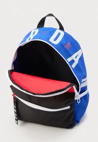 Μπλε και μαύρο σακίδιο με λευκά και κόκκινα γράμματα, μπροστινή τσέπη με φερμουάρ, το κυρίως διαμέρισμα ανοιχτό που δείχνει την κόκκινη εσωτερική πτέρυγα.