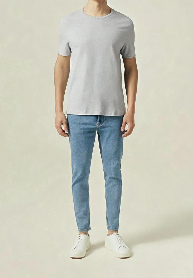 Mężczyzna w jasnoszarej koszulce z krótkim rękawem, jasnoniebieskich skinny jeansach i białych trampkach, stojący na jednolitym beżowym tle.