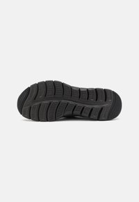 Black athletic shoe sole with a textured rubber surface, featuring deep grooves and a rounded heel for traction and stability.