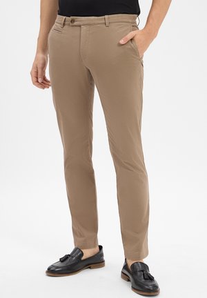Mann in schmal geschnittenen beigen Hosen und schwarzen Leder-Tassel-Loafers, stehend mit einer Hand in der Tasche vor einem weißen Hintergrund.