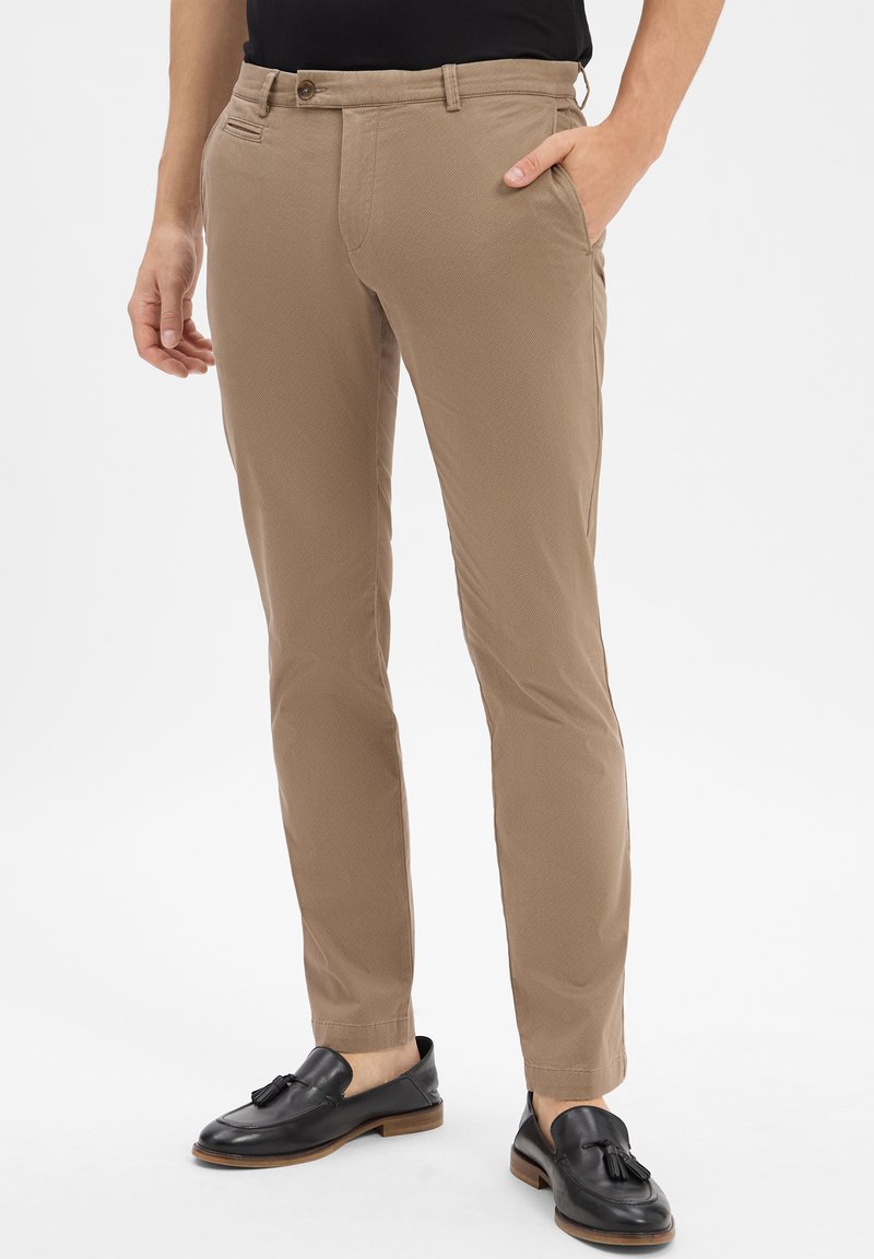 Mężczyzna w dopasowanych beżowych spodniach i czarnych skórzanych loafersach z frędzlami, stojący z jedną ręką w kieszeni na białym tle.