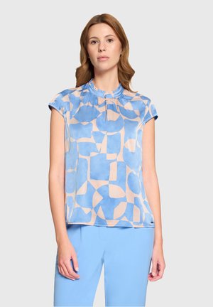 Woman with light brown hair wearing a short-sleeve blue and peach geometric patterned blouse and light blue pants, standing against a plain background.