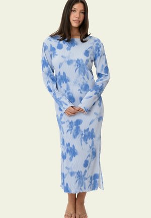 Frau trägt ein midilanges Kleid mit langen Ärmeln und blau-weißem Blumenmuster, steht mit verschränkten Händen vor einem schlichten hellen Hintergrund.