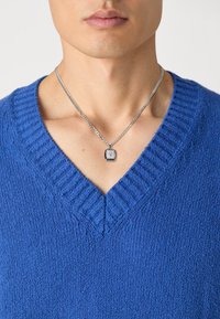 Pull en V en tricot texturé bleu vif. Un collier en chaîne argentée avec un pendentif carré est suspendu au cou. Fini lisse et soigné.