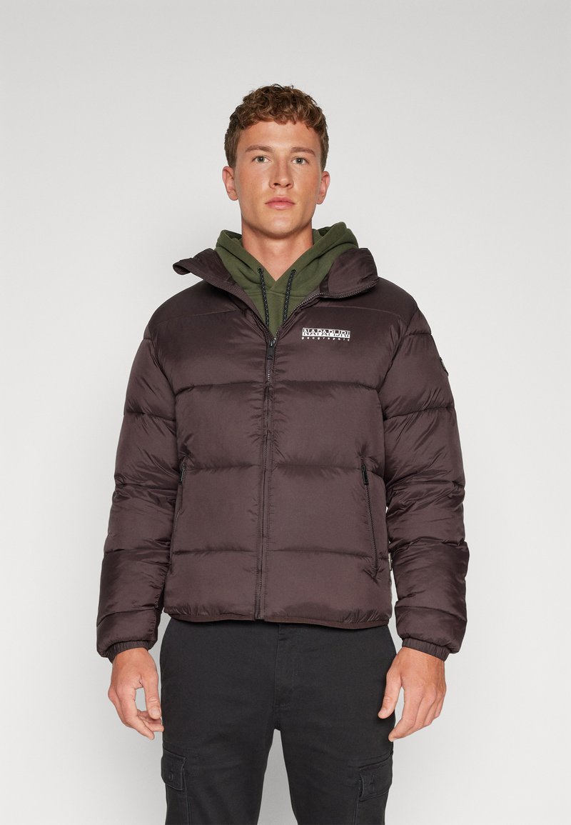 Mörkbrun puffjacka med huva, dragkedjestängning, quiltad textur och logodetalj. Bärs över en grön hoodie och kombineras med svarta byxor.