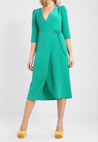 Green wrap dress with three-quarter sleeves, a deep V-neckline, tie belt at the waist, and a smooth, flowing fabric. Paired with yellow sandals.