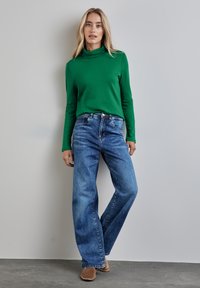 Grüner Baumwollrollkragenpullover kombiniert mit hellblauen, hochgeschnittenen, weit geschnittenen Jeans. Das Model steht auf einer grauen Fläche vor einem schlichten Hintergrund.