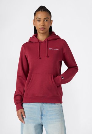 Vrouw draagt een bordeauxrode Champion hoodie met voorzak en lichtblauwe spijkerbroek, staand tegen een effen witte achtergrond.