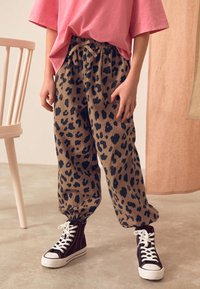 Leopard print jogger pants in brown with dark spots, featuring an elastic waistband and cuffed ankles, paired with black high-top sneakers.