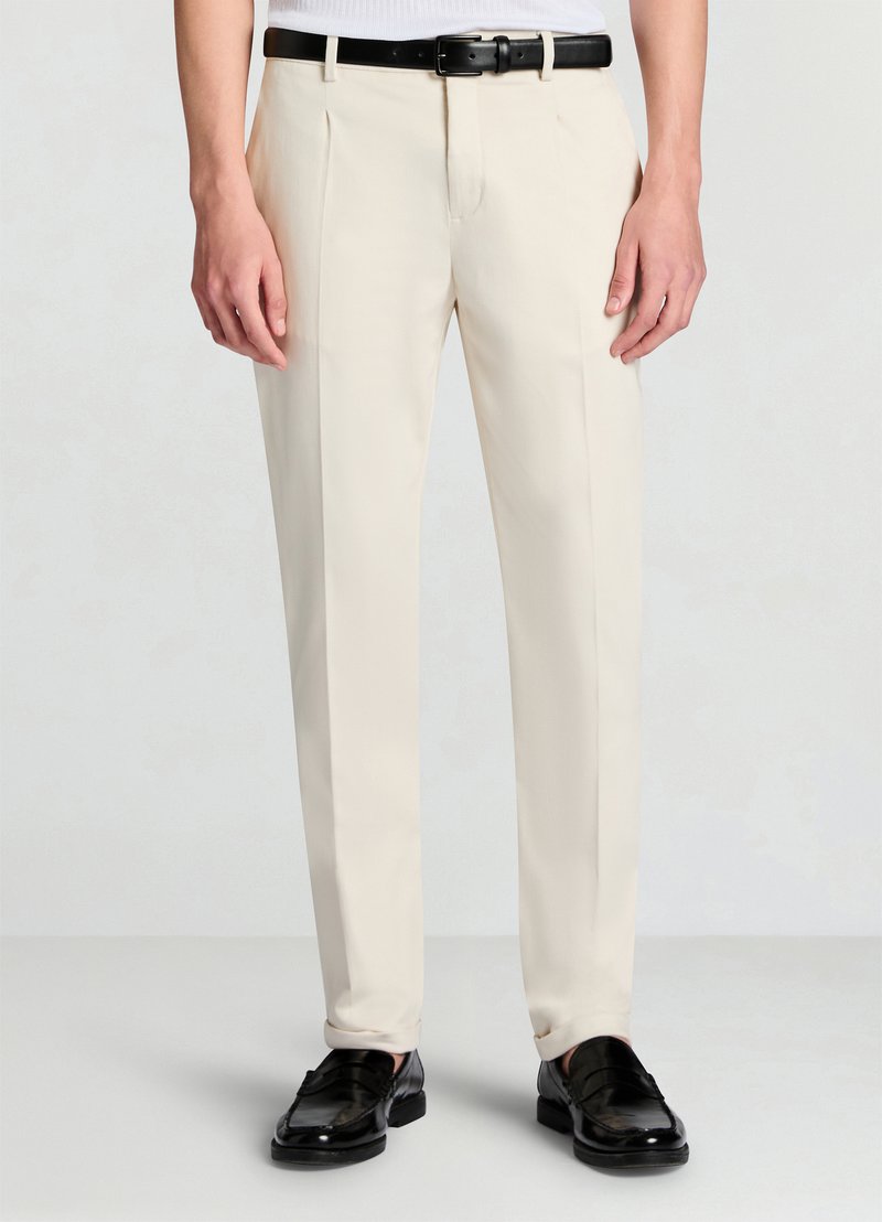 Pantaloni beige da donna con taglio dritto, fronte piatto, due pieghe e cintura in pelle nera. Indossati con mocassini neri. Tessuto liscio.