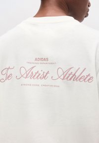 Άτομο που φοράει λευκό μπλουζάκι Adidas με ροζ κείμενο στην πλάτη που λέει "The Artist Athlete" και "Strong core. Creative mind."