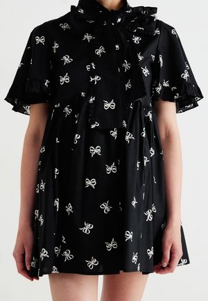 Schwarzes Kleid mit weißen Schleifenmustern, flatternden Ärmeln und einer großen, gerafften Schleife am Ausschnitt, getragen von einer Person vom Hals bis zur Mitte des Oberschenkels.