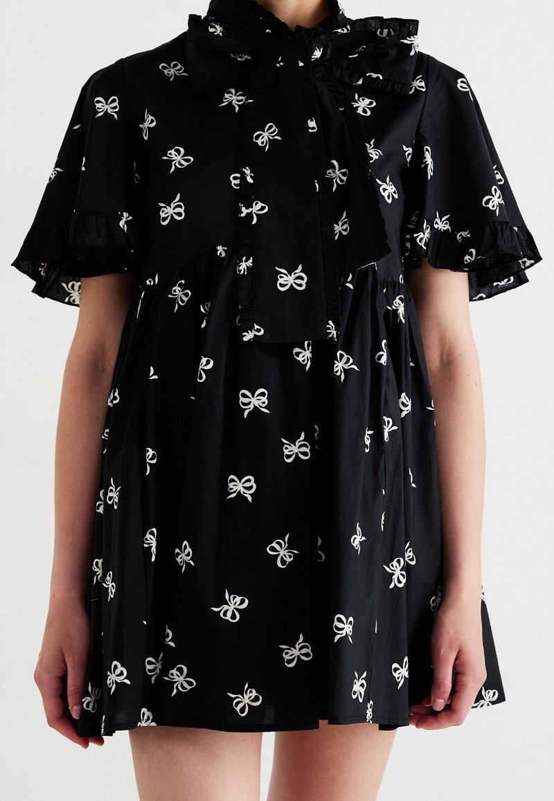 Robe noire avec motifs de nœuds blancs, manches volantées et un grand nœud volanté attaché au niveau du col, portée par une personne du cou jusqu'au milieu des cuisses.