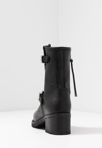Černá kožená kotníková bota s texturovaným povrchem, dvěma stříbrnými přezkami a krátkým klínovým podpatkem. Obsahuje zip a poutko.