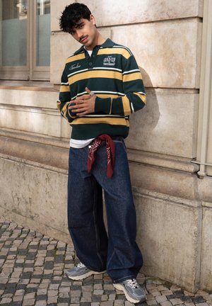 Ung mand med krøllet hår læner sig mod en stenmursvæg iført en stribet grøn og gul sweater, posede jeans, sneakers og et rødt bandana om livet.