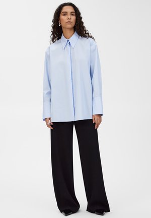 Kobieta o kręconych włosach, ubrana w luźną jasnoniebieską koszulę z guzikami i szerokim kołnierzem oraz czarne spodnie o szerokich nogawkach, stojąca na białym tle.
