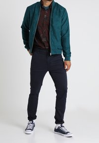 Teal, zip-up bomber jacket in soft material, paired with a dark patterned shirt, dark pants, and navy sneakers. Casual, fitted design.