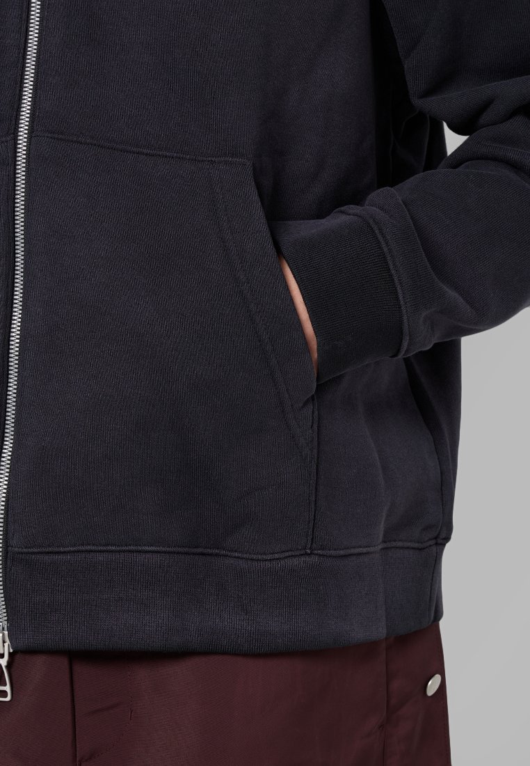 Dunkelgrauer Kapuzenpullover mit Reißverschluss, strukturierter Stoff, seitlichen Taschen und gerippten Bündchen, ergänzt durch kontrastierende weinrote Hose.