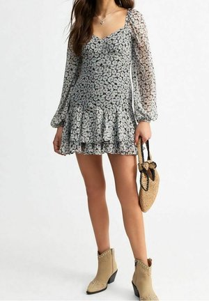 Kobieta ubrana w długą sukienkę w czarno-białe kwiaty z falbanową spódnicą, beżowe botki z ćwiekami i trzymająca plecioną torebkę.