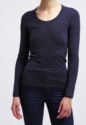 Persona che indossa una camicia aderente a maniche lunghe blu scuro e pantaloni blu scuro abbinati contro uno sfondo chiaro e uniforme.