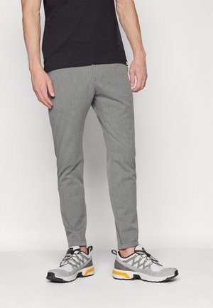 Man draagt slanke grijze broek, een zwart shirt en grijs-witte sneakers met gele accenten, staand tegen een witte achtergrond.