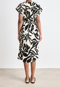 Femme portant une robe mi-longue à imprimé abstrait de feuilles noir et blanc, debout pieds nus sur un sol clair contre un fond uni.