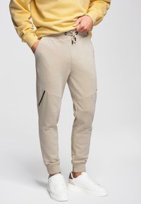 Homme portant un pantalon de jogging beige avec des détails de fermeture éclair noirs et des baskets blanches, debout avec une main dans la poche et un sweat-shirt jaune.