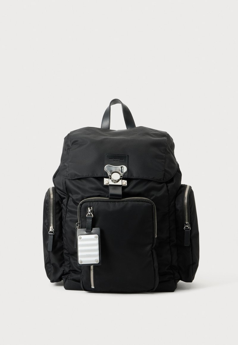 Sac à dos noir avec des poches zippées à l'avant et sur les côtés, une boucle argentée sur le rabat supérieur et une petite étiquette blanche suspendue à la fermeture éclair de la poche avant.
