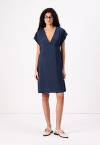 Rochie bleumarin cu mânecă scurtă, de tip V, dintr-un material fluid, cu o lungime până la genunchi și textură minimă. Asortată cu pantofi slip-on de culoare deschisă.