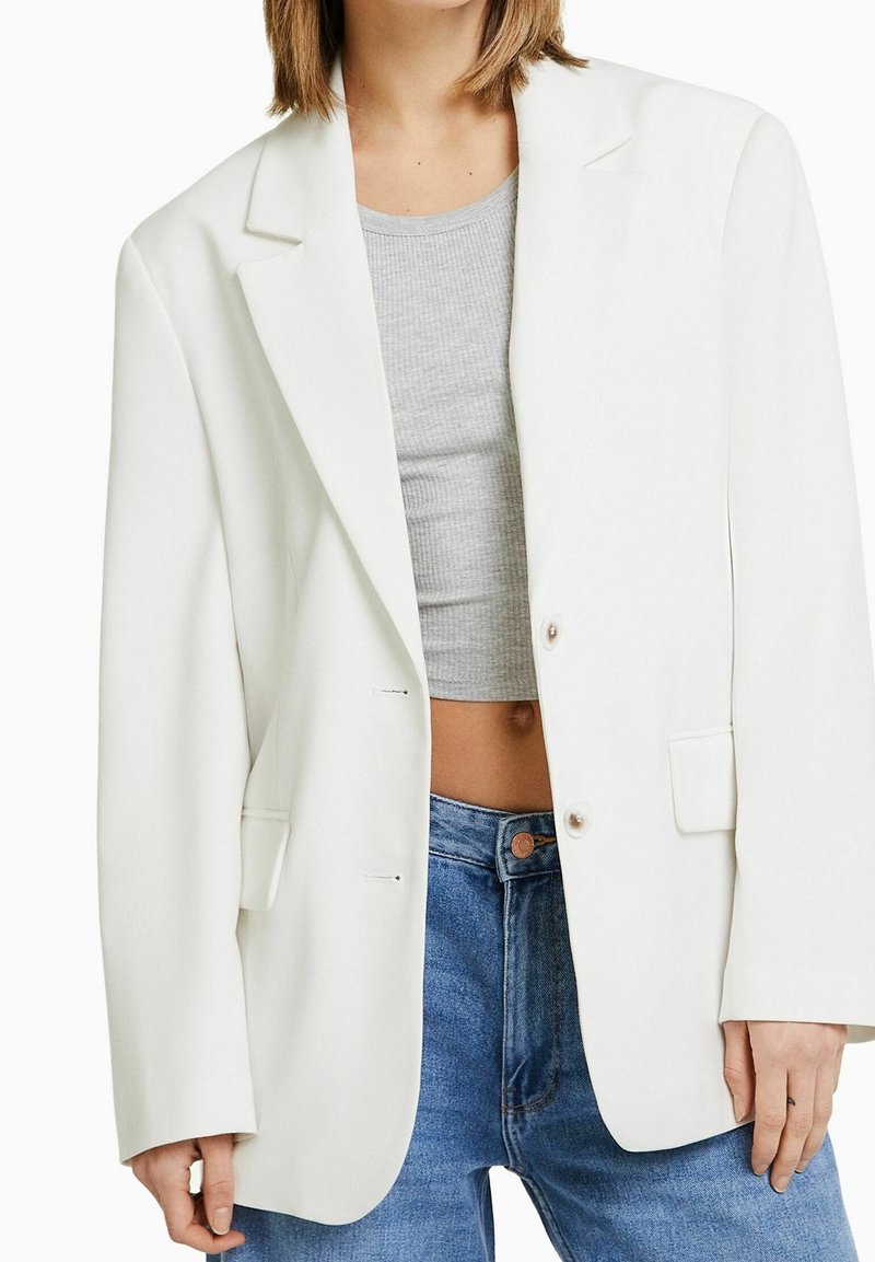 Femme portant un blazer blanc oversize par-dessus un top court gris et un jean bleu, debout les mains relâchées sur les côtés.