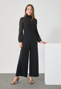 Black jumpsuit with a high neck and sheer puff sleeves; wide-leg design. Features a seamless waist and accented with strappy black heels.