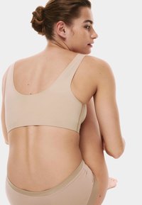 Mujer joven con el cabello recogido en un moño, vistiendo un sujetador y ropa interior beige sin costuras, sentada con una rodilla levantada sobre un fondo blanco.