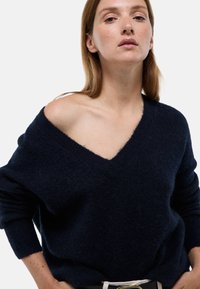 Pull en maille marin avec un décolleté en V plongeant et une coupe oversized. Il présente des manches longues et un matériau doux et texturé, mettant en avant un style décontracté.