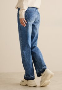 Blaue Jeans aus Denim mit entspannter Passform, klassischem Fünf-Taschen-Design und subtiler Ausbleichung. Getragen mit weißen Sneakern.