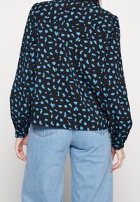 Camicia nera con motivo a pois blu, maniche lunghe e colletto. Il tessuto ha un aspetto testurizzato. Abbinata a jeans blu chiaro.