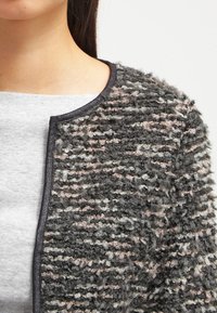 Texturerad grå jacka med rosa och vita prickar, rund hals och svart kant. Tyget verkar mjukt och fluffigt med en lagerdesign.