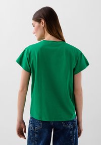 T-shirt verde in cotone con maniche corte, scollatura rotonda e vestibilità rilassata. Il tessuto appare liscio con un semplice design sul retro.