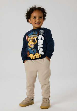 Smiling young boy with curly hair wearing navy Paw Patrol Chase sweatshirt, beige pants, and beige shoes, hands in pockets, on light gray background.