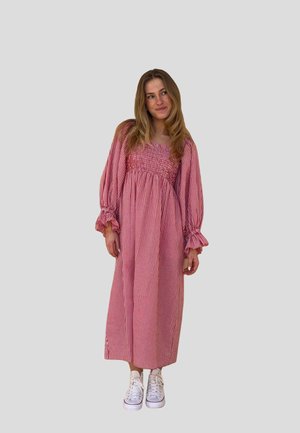 Kvinne med langt hår som har på seg en rød og hvit rutete lang kjole og hvite joggesko, står og ser til siden.