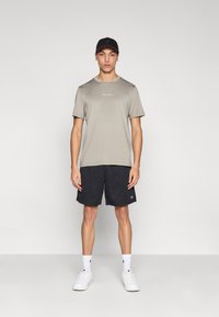 Ανοιχτό γκρι πουκάμισο με κοντά μανίκια και μικρό λογότυπο, συνδυασμένο με μαύρο σορτς από δίχτυ και άσπρα αθλητικά παπούτσια. Μαύρο καπέλο ολοκληρώνει την εμφάνιση.