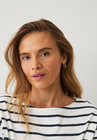 Gestreifte Baumwollbluse in Weiß mit dunkelblauen horizontalen Streifen, ausgestattet mit einem weiten Ausschnitt. Haare in sanften Wellen gestylt, Hoops als Ohrringe getragen.