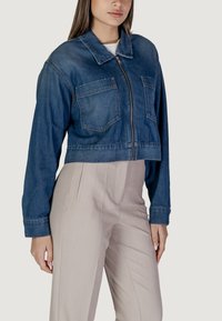 Giacca di jeans cropped in blu scuro, con due tasche sul petto e zip frontale. Abbinata a pantaloni beige chiaro sartoriali.