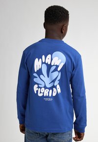 Langärmliges blaues T-Shirt mit einem großen weißen und hellblauen Grafik auf der Rückseite, die den Text "MIAMI FLORIDA" und ein kreisförmiges Detail zeigt.