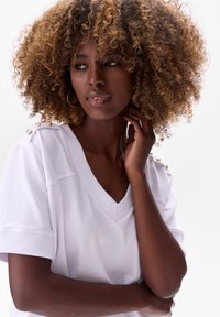 Witte V-hals top met korte mouwen, voorzien van gouden accentknopen op de schouders. Zachte stof, gladde textuur, aansluitend ontwerp.