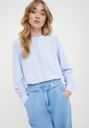 Helle blaue Bluse mit langen Ärmeln aus strukturiertem Stoff, mit Spitzenverzierungen an der Vorderseite und rundem Ausschnitt, kombiniert mit hoch taillierten Jeanshosen.