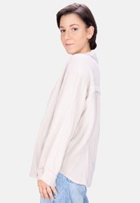 Light beige long-sleeve shirt with a textured fabric and button cuff, paired with light blue jeans. Features a relaxed fit and subtle details.