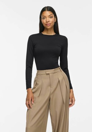 Femme aux longs cheveux foncés portant un haut noir cintré à manches longues et un pantalon beige taille haute à jambes larges, debout sur un fond blanc.